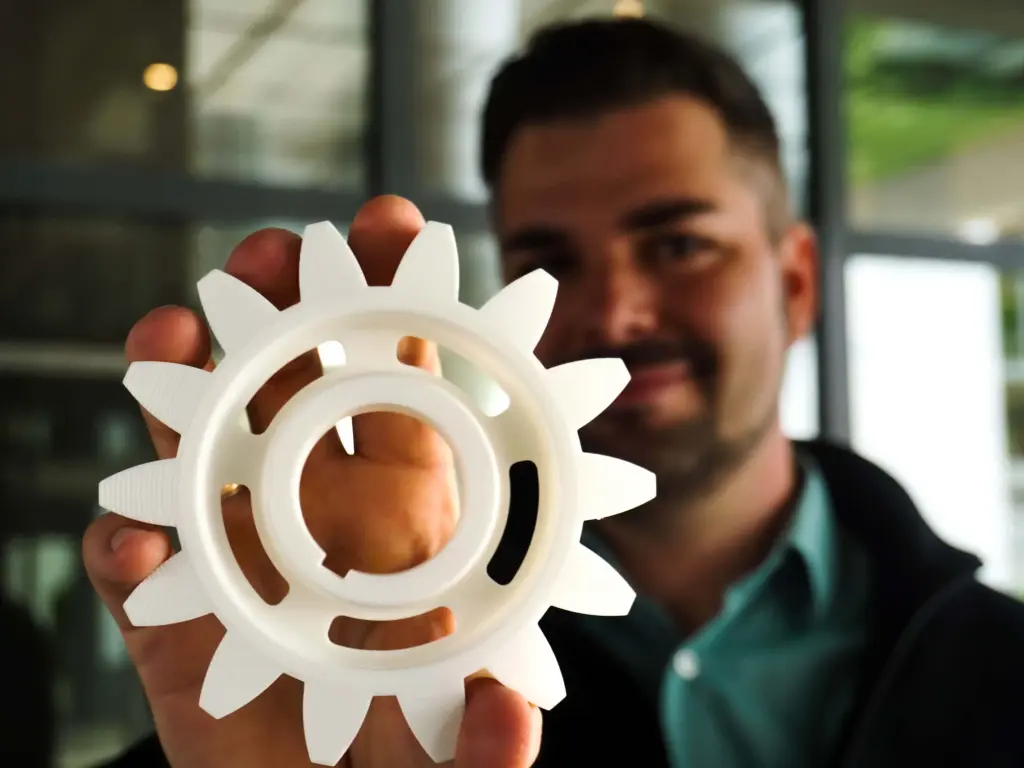 Ein Mann, der Gründer von Revoneer, Dominik Blendl hält ein weißes mit einem 3D-Drucker gedrucktes Kunststoffzahnrad in die Höhe, symbolisiert technische Beratung und Erstberatung.