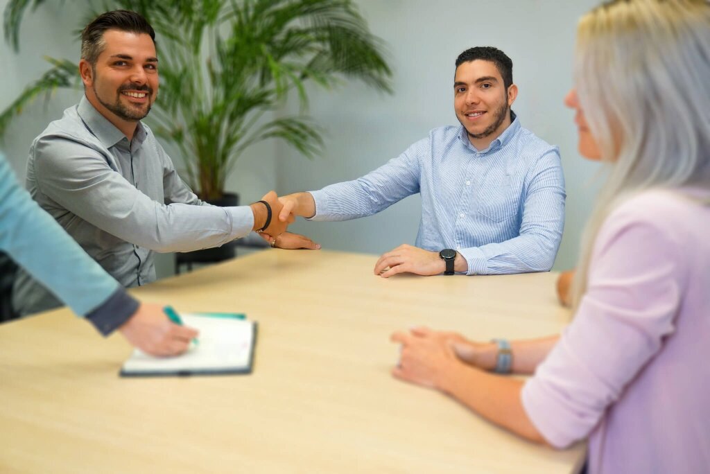 Zwei Männer lächeln in die Kamera und schütteln sich die Hände an einem Besprechungstisch während eines geschäftlichen Meetings.